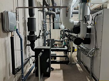Technikraum mit gedämmten Rohrleitungen, Ventilen und technischen Anlagen an einer Wand.